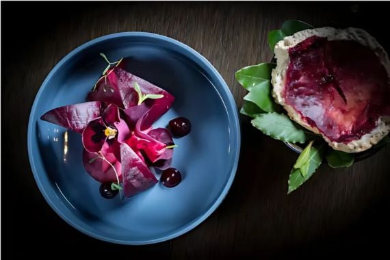 Elegant plate of vibrant red beetroot garnished with microgreens and cherries, paired with a rustic side of leafy greens.
