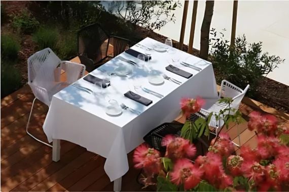 An elegant outdoor dining setup with a white tablecloth and minimalist tableware, surrounded by greenery and pink blossoms.