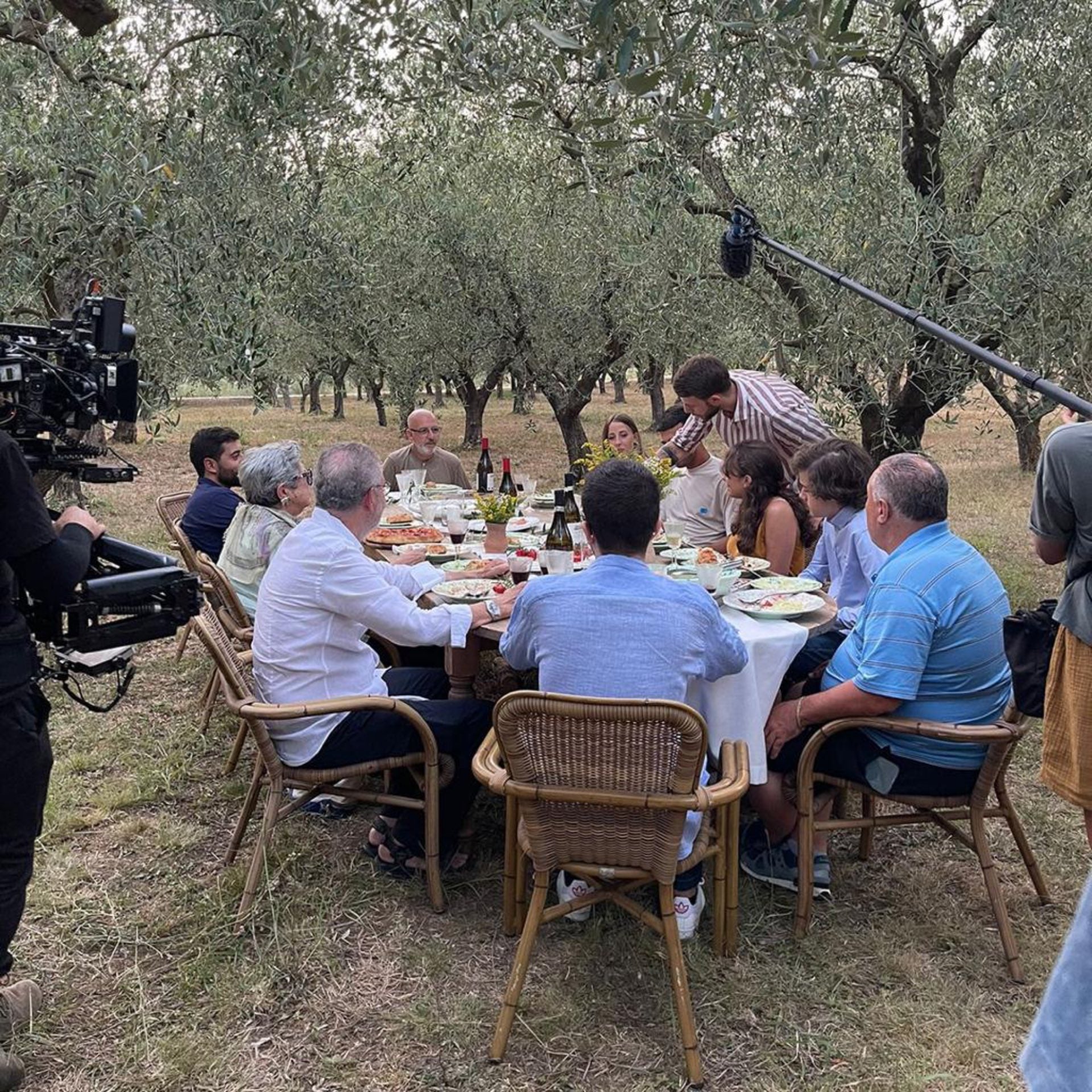 a group of diners seated around a table in an olive grove being filmed