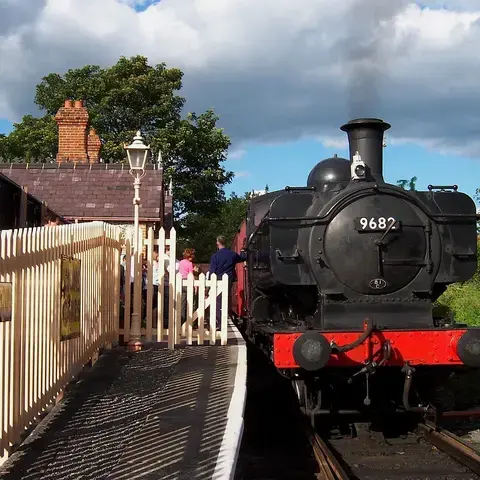 Vintage black steam locomotive number 9682, resting at a quaint station with white fences, under a partly cloudy blue sky