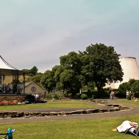 Outdoor concert in a park with a gazebo, audience seated on the grass, and trees surrounding the scene on a sunny day