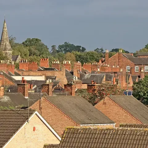 Historic town rooftops with red brick chimneys, a stone church spire in the background, and lush greenery on the horizon.