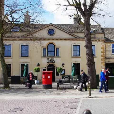 Historic building with arched windows, ‘Bill’s’ sign, red postbox, trees, and pedestrians on a busy street