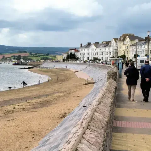 Beachfront promenade with walkers, a sandy beach, and rows of Victorian houses under a cloudy sky
