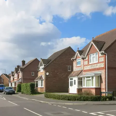 Suburban street with rows of red-brick houses, parked cars, and a partly cloudy blue sky