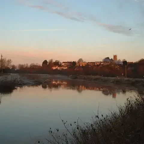Sunset view of a riverside village with a church tower, reflecting in the calm water under a pastel sky.