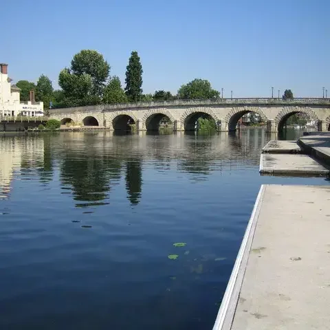 Arched stone bridge spanning a calm river with lily pads, riverside buildings, and clear blue sky