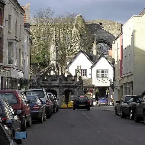 Historic town center with parked cars, medieval market cross, and ancient ruins in the background under a partly cloudy sky