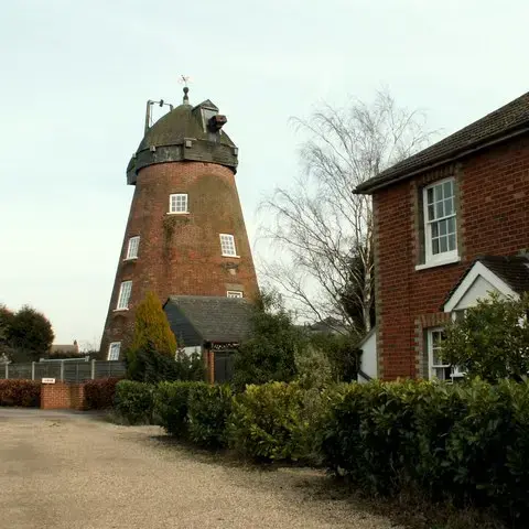 Historic windmill with a red brick base, surrounded by greenery and traditional houses, under a clear sky
