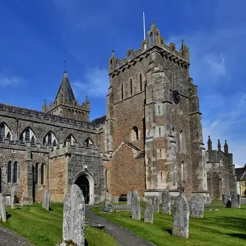 Historic parish church with Gothic architecture, a tall clock tower, and a graveyard under a bright blue sky.
