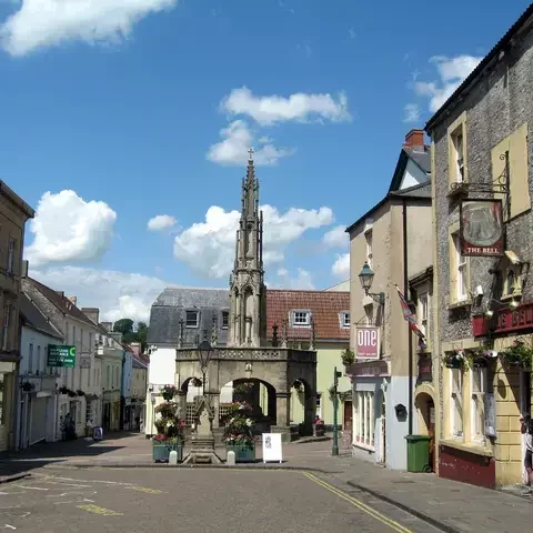 Sunny market square in a historic town with The Bell pub, shops, and a decorative stone monument under a blue sky with clouds