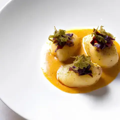 3 delicate scallops on a rich orange sauce, topped with finely chopped greens and purple garnish, arranged in a white bowl