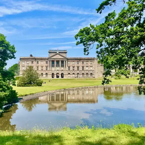 Elegant mansion with a grand facade reflected in a calm pond, surrounded by lush greenery under a blue sky