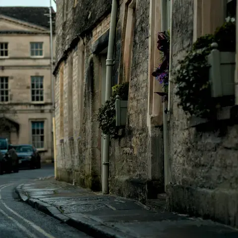 Quaint stone street with flower-filled window boxes,framed by historic architecture and parked cars under a soft,muted light