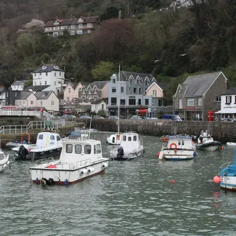 Small harbor with fishing boats, colorful houses, and lush hills in the background on a cloudy day.