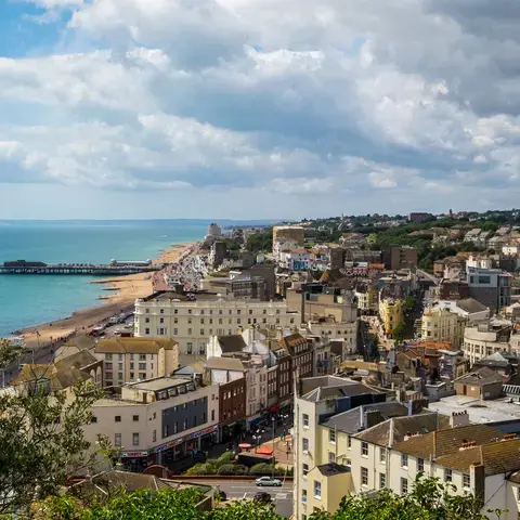 Coastal cityscape with a sandy beach, pier, and vibrant buildings overlooking the turquoise sea under a partly cloudy sky.