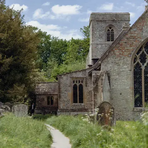 Stone church in countryside, surrounded by grass and gravestones, with trees and blue sky in the background