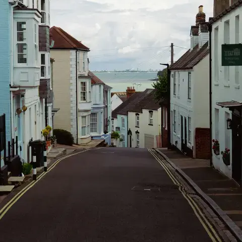 A narrow coastal street lined with pastel-colored houses leading to a serene view of the sea under a cloudy sky.
