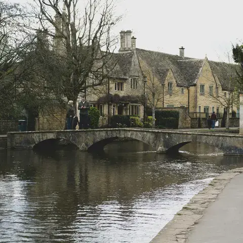 A stone bridge arches over a tranquil stream in a quaint village, flanked by charming cottages and budding spring trees