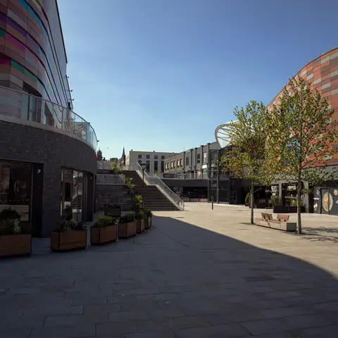 Modern city square with colorful architecture, glass-fronted buildings, stairs, trees, and restaurants under a clear blue sky