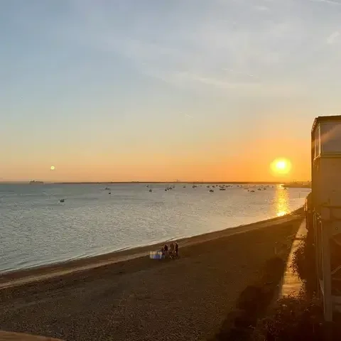 Golden sunset over a calm sea, with small boats scattered on the horizon and silhouettes of people on a sandy beach