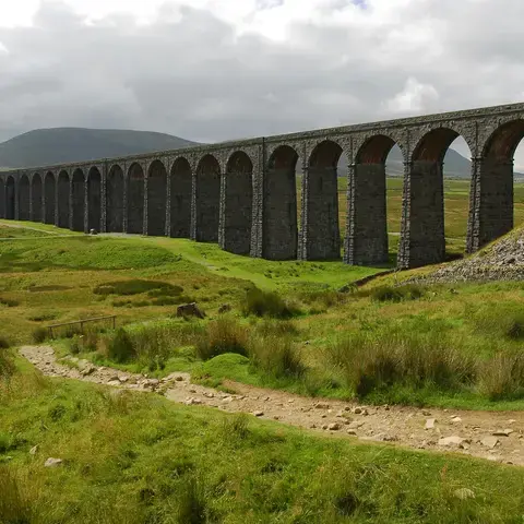 Stone viaduct with arches spans green moorland under cloudy skies, a dirt path winds towards the iconic structure