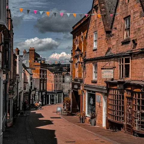 Charming old town street with antique shops, bunting overhead, and a backdrop of scenic countryside under a partly cloudy sky