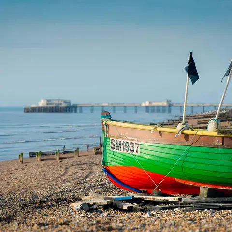 Vibrant fishing boat rests on a pebbled beach with a calm sea and distant pier, capturing a serene coastal morning scene