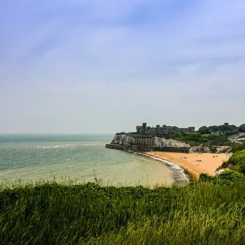 Coastal view with sandy beach, cliffs, and historic building overlooking calm sea under a hazy sky