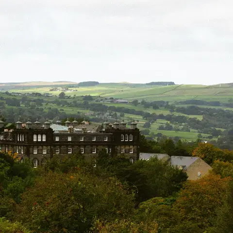 Historic stone building nestled in lush greenery, overlooking the rolling countryside of Yorkshire under a cloudy sky