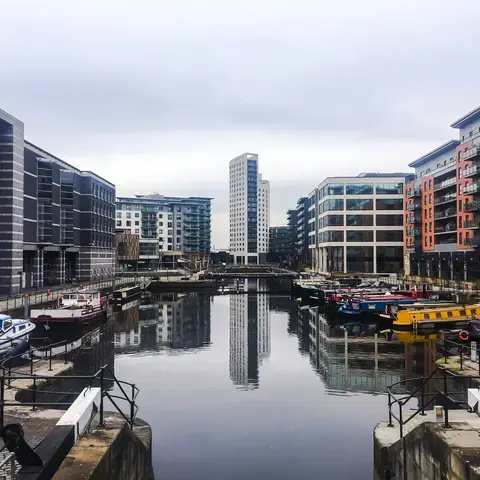 Canal boats docked along a calm waterway, framed by modern buildings under a cloudy sky. A peaceful urban scene