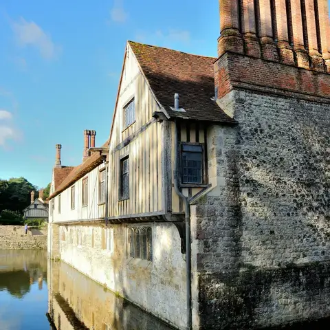 Tudor-style building with timber framing and tall brick chimneys reflecting in a calm moat under a blue sky