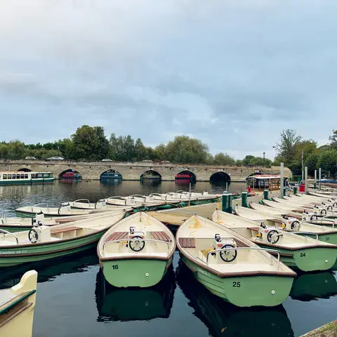 Cream and green boats docked near a stone bridge, reflecting on calm waters under a cloudy sky with trees in the background