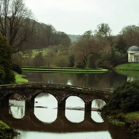 Stone bridge over a serene lake with a domed temple in the background, surrounded by lush greenery and peaceful scenery