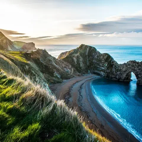 Scenic coastal view of a natural rock arch over blue water, with grassy cliffs under a bright sky.