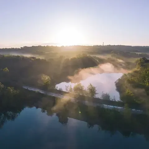 Serene sunrise over a misty forest with a reflective lake and a winding road cutting through the lush greenery.