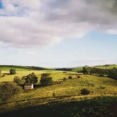 Rolling green hills with a lone stone barn, scattered sheep, and sunlight breaking through a partly cloudy sky