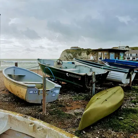 Weathered fishing boats rest on a pebbled shore under a cloudy sky, with cliffs and a calm sea in the background