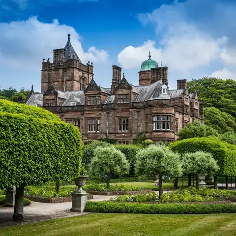 Historic brick manor surrounded by manicured gardens, green hedges, and a bright blue sky with scattered clouds.