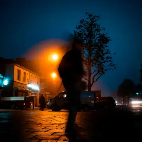 Nighttime city street with glowing streetlights, a silhouetted figure in motion, and storefronts illuminated by neon signs