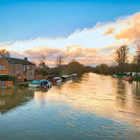 Golden-hour view of a serene river lined with boats, brick houses, and trees,reflecting the vibrant colors of the setting sun