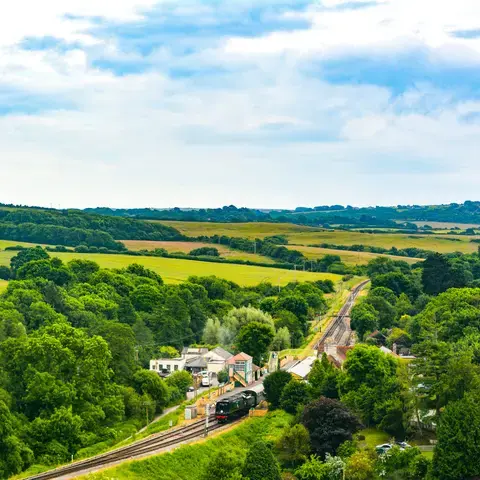 Scenic countryside with green fields, trees, and a small train station nestled between railway tracks under a cloudy sky