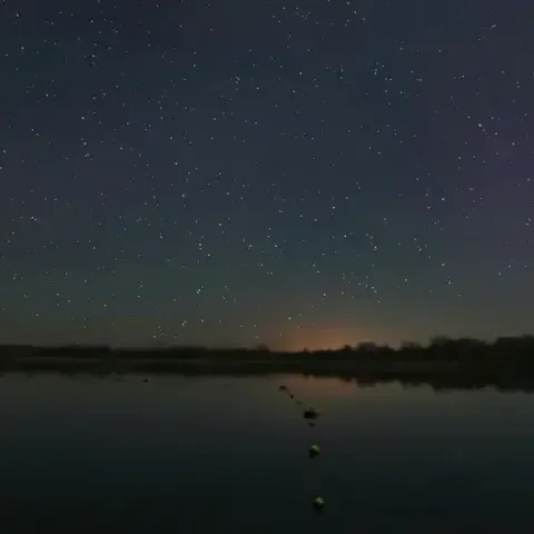 Starry night sky over a calm lake, faint orange glow on the horizon, and a serene atmosphere perfect for stargazing