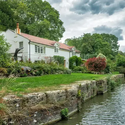 A charming countryside home with a red-tiled roof, lush gardens, and a tranquil riverside setting under a cloudy sky