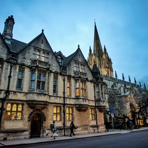 Gothic-style building illuminated at dusk with pointed spires, arched windows, and pedestrians walking along the street