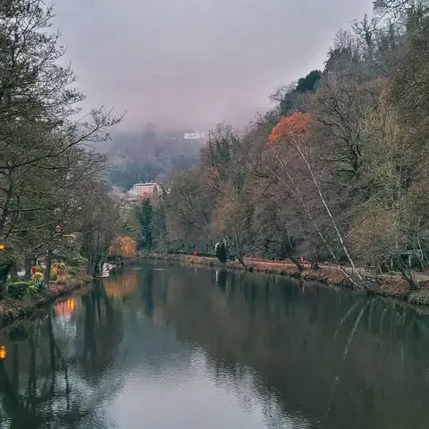 Serene river flanked by bare winter trees and a foggy hillside in the distance, with paths and benches lining the riverbanks