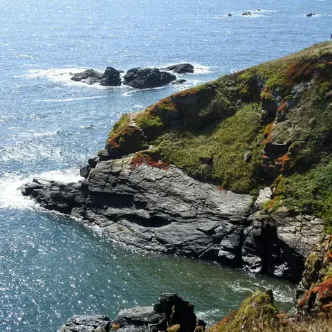 Rocky coastal cliff with green vegetation overlooking the ocean on a sunny day, with people standing on a lookout point in th