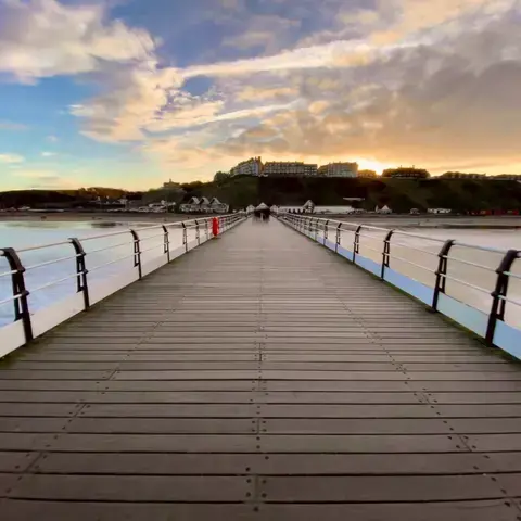 Wooden pier extending towards the horizon, flanked by calm waters and hills, with a glowing sunset illuminating the sky