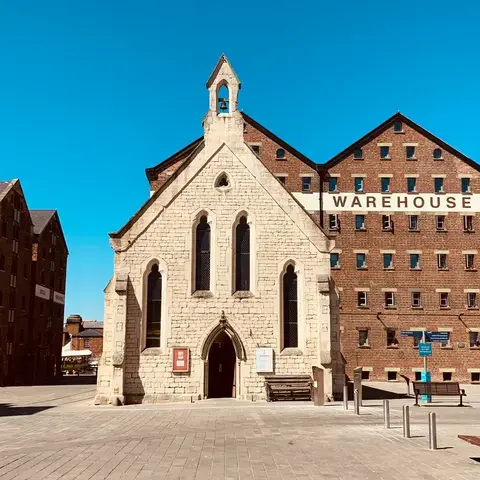 Historic stone chapel surrounded by red-brick warehouses under a bright blue sky, blending tradition with industrial heritage