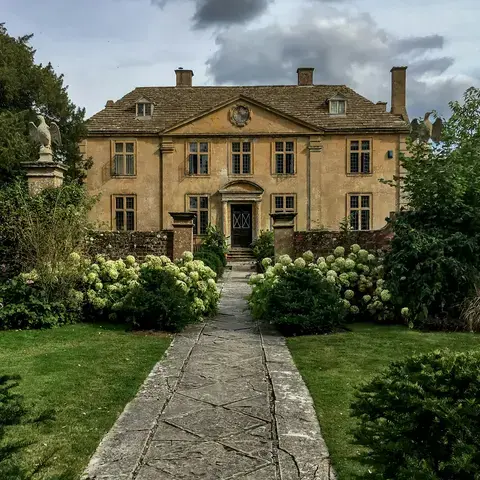 Elegant historic manor house with a stone pathway, surrounded by lush gardens and hedges under a cloudy sky